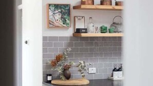 Una cocina con encimera oscura, tres taburetes, estante colgante con utensilios de cocina y salpicadero de azulejos.