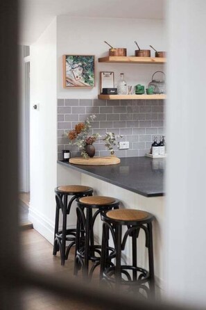 Eine Küche mit dunkler Arbeitsplatte, drei Hocker, einem hängenden Küchenregal und einer gefliesten Spritzschutzwand.