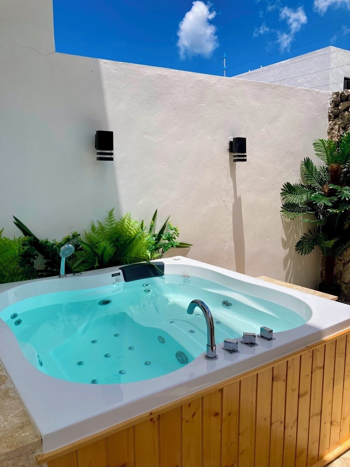 A hot tub with a wooden deck, surrounded by greenery and a white wall.