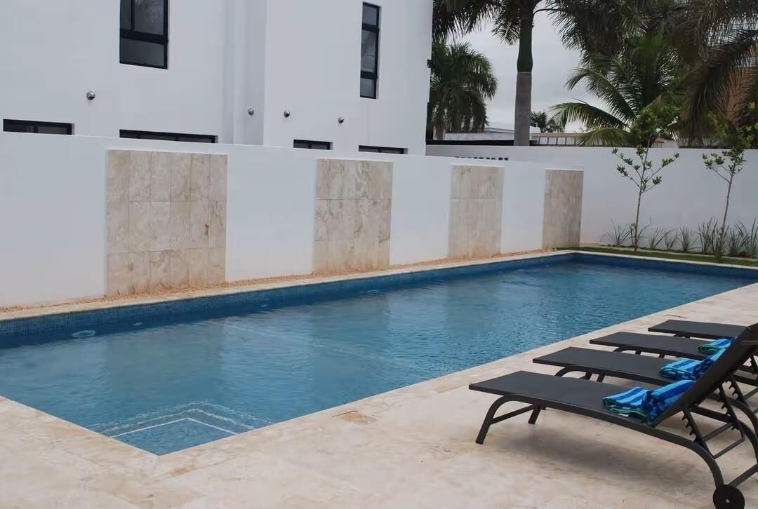 Ein Swimmingpool mit gefliestem Rand, eine weiße Wand mit Fenstern und einige Liegestühle.