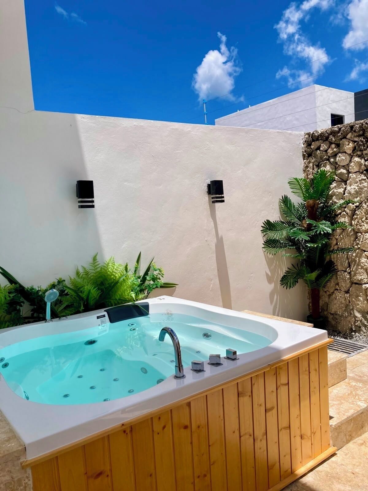 Un bain à remous doté d’une terrasse en bois, entouré de plantes et d’un mur de pierre, sous un ciel bleu dégagé.