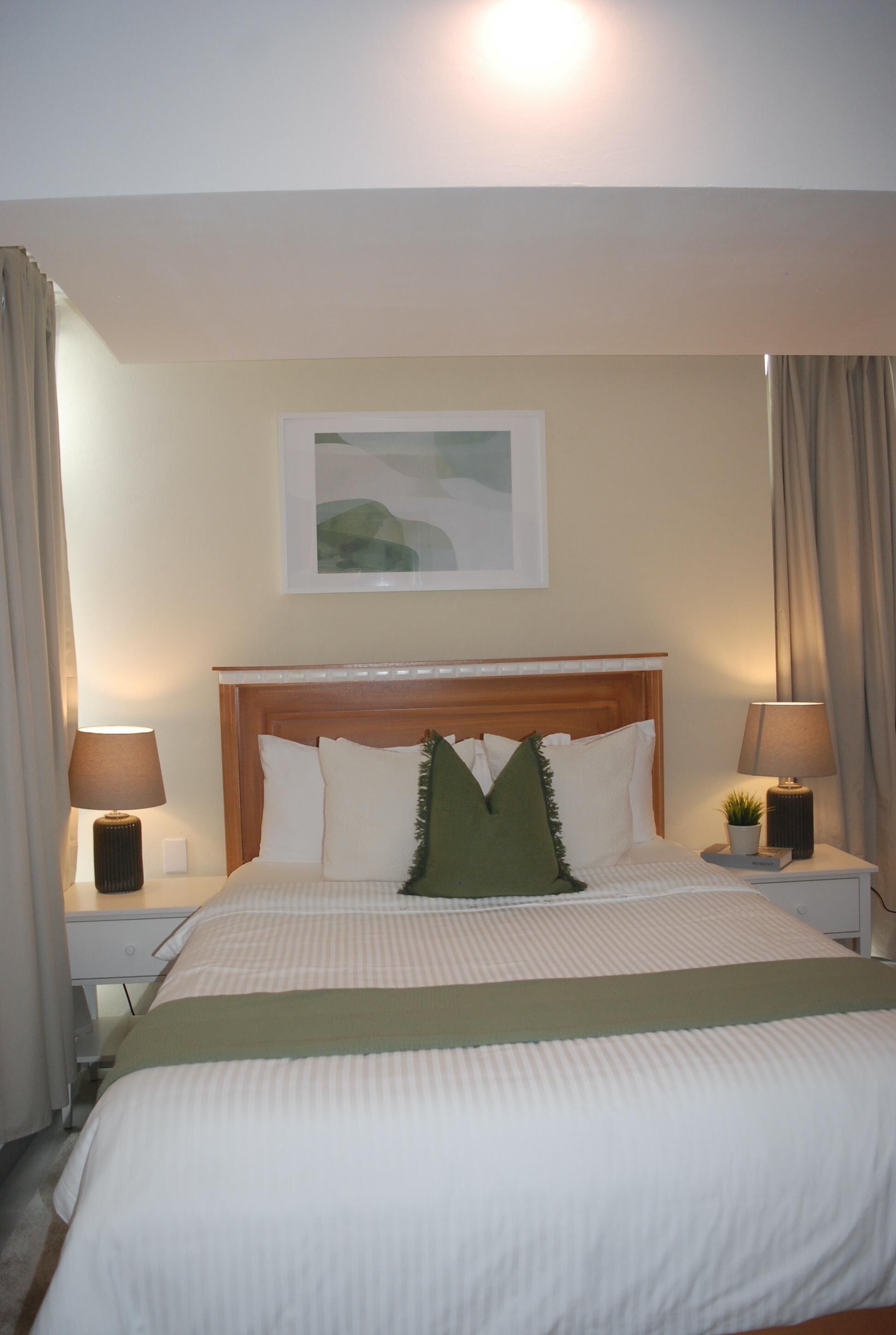 A hotel room with a bed, two bedside lamps, a nightstand, a framed picture on the wall, and a window with curtains.