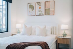 Une chambre d’hôtel comprenant un lit, deux tables de chevet avec des lampes, une fenêtre et trois œuvres d’art encadrées accrochées au mur.