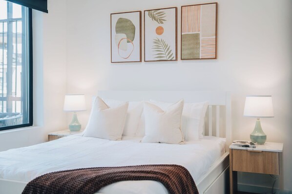 Ein Hotelzimmer mit einem Bett, zwei Nachttischen mit Lampen, einem Fenster und drei gerahmten Kunstwerken an der Wand.