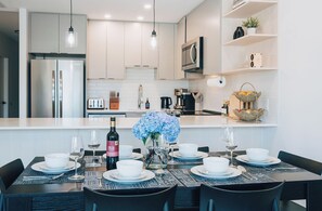 A modern kitchen with white cabinetry, stainless steel appliances, and a dining area set for six with a table set for a meal. - TNSKI - Modern mountain retreat with hot tub and stunning views (Revelstoke)