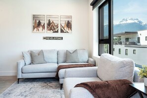 Ein modernes Wohnzimmer mit einer grauen Eckcouch, zwei gemusterten Zierkissen, einer braunen Decke und einem großen Fenster mit Blick auf die Berge.