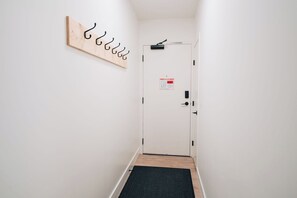 Un couloir avec un porte-manteau en bois, une porte ornée d’un panneau indiquant les consignes de sécurité incendie et un paillasson noir à l’entrée.