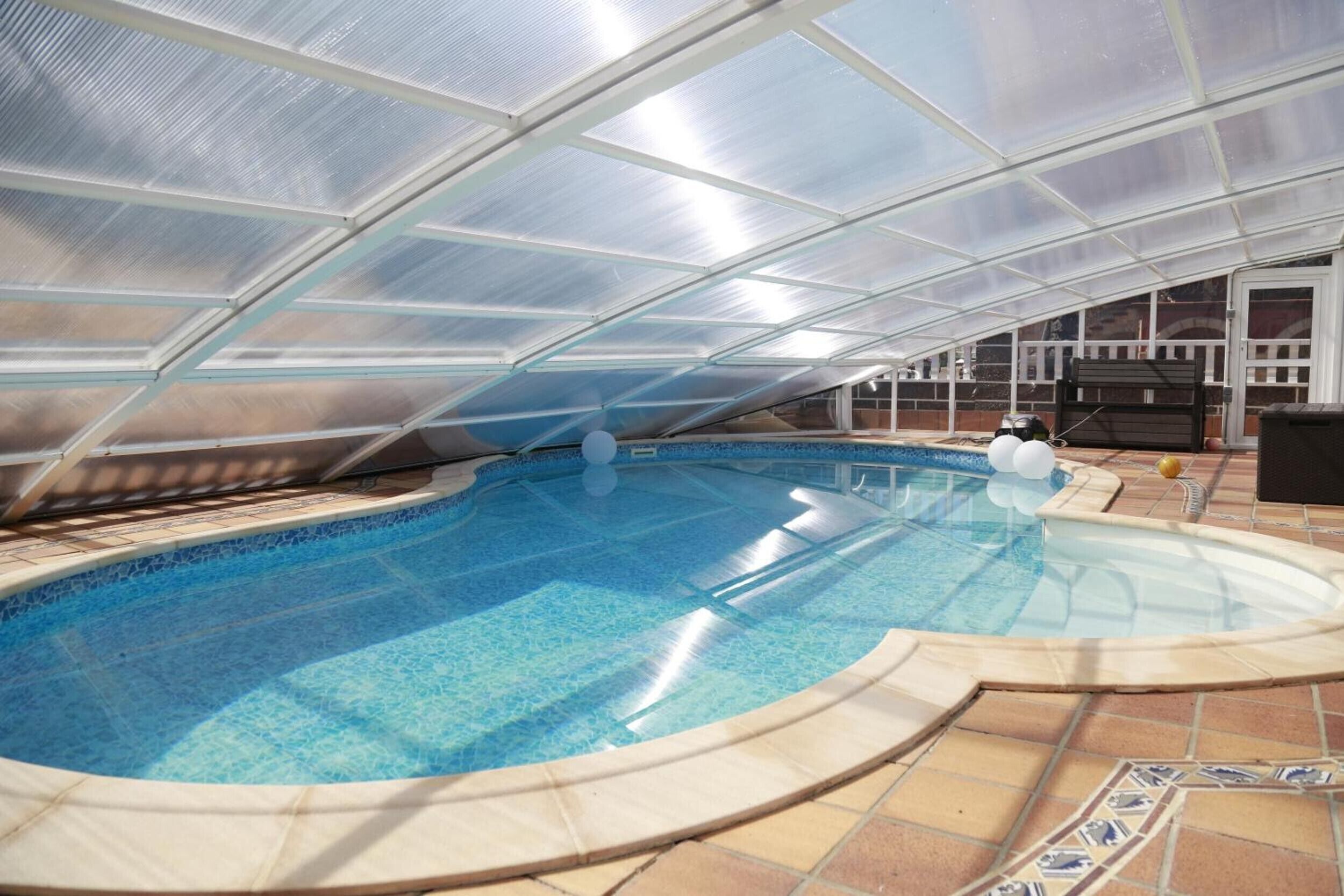 A swimming pool with a glass ceiling structure, surrounded by a tiled deck.