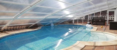 A swimming pool with a glass ceiling structure, surrounded by a tiled deck.