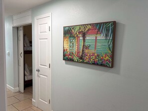 Ein Flur mit einer Tür zu einem Zimmer, das ein Bett und einen Kleiderschrank enth ält. An der Wand hängt ein Bild mit einer tropischen Szene.