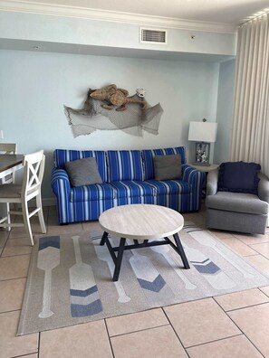 A living room with a striped sofa, a round coffee table, and a dining area with a chair and table. - Gulf Front Condo with Pool, Hot Tub & Beach Access (Panama City Beach)