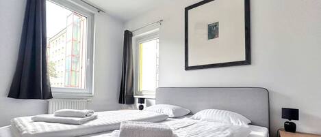Una habitación de hotel con una cama, dos ventanas, un cuadro enmarcado, una mesita de noche y una lámpara.