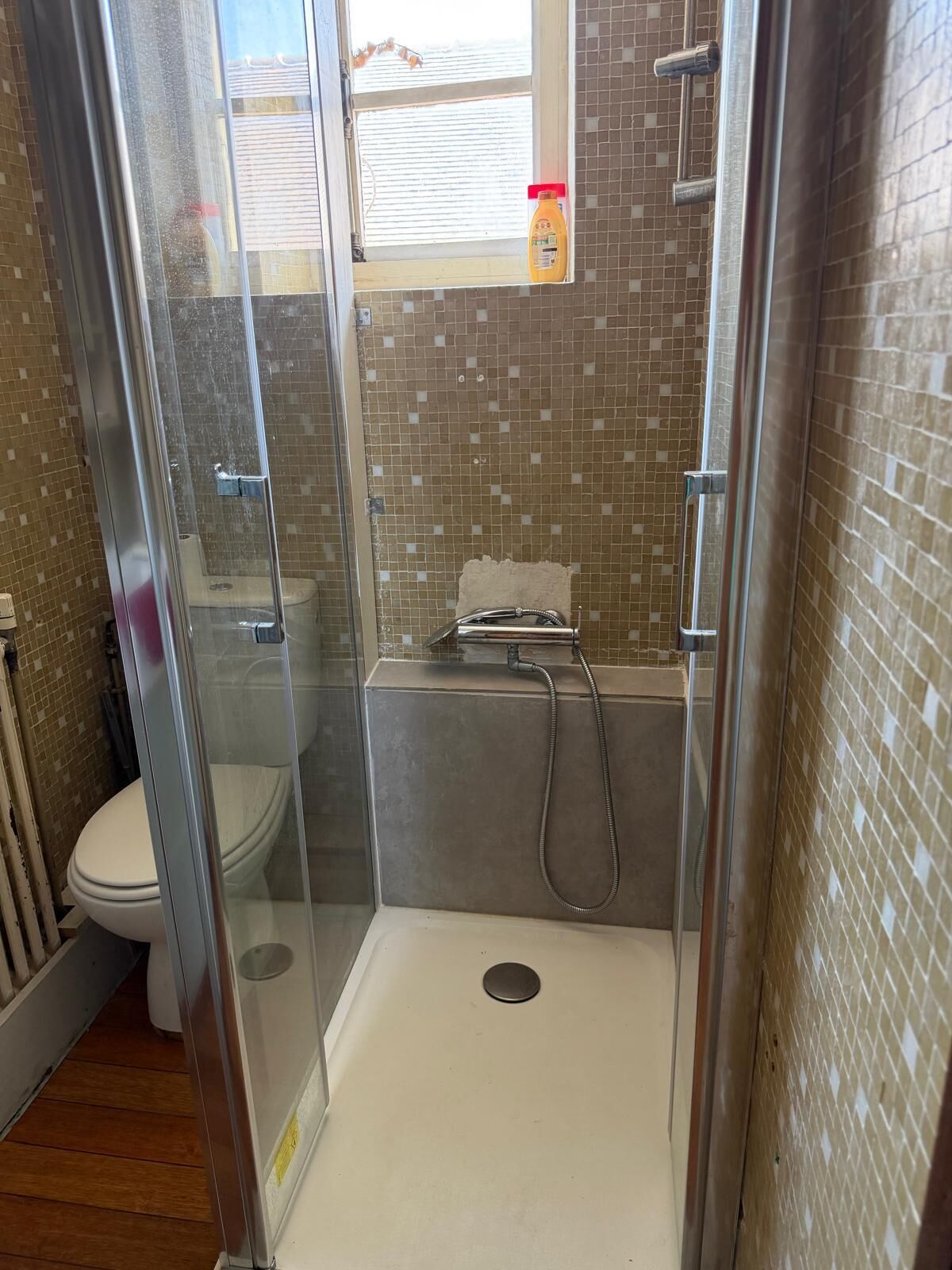 Ein Badezimmer mit einer gläsernen Duschkabine, einer Toilette und einem Fenster mit einem Reinigungsmittel auf der Fensterbank.