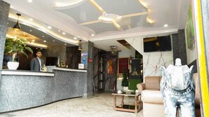 Zona de recepción de un hotel con mostrador de mármol, escritorio, televisor montado en la pared, una estatua e iluminación empotrada en el techo.