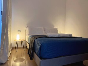 A single bed with a blue bedspread, a bedside table with a lamp, and a curtain on the left. - Atlantico Apartment: Air Conditioning, Swimming Pool (Santa Cruz de Tenerife)