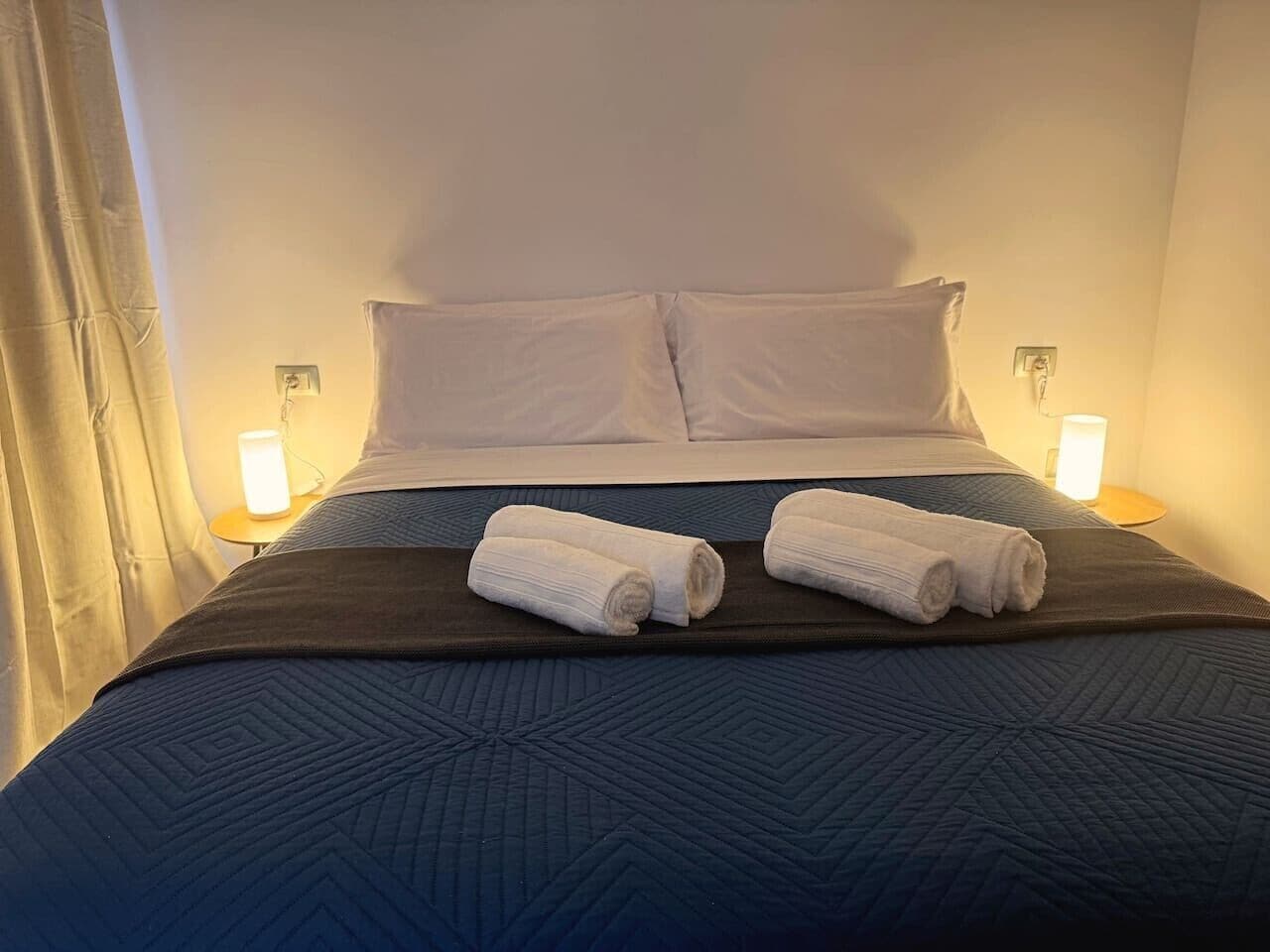 A hotel room with a bed, two white pillows, a dark blue quilted bedspread, and two rolled towels on a tray.