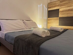 Ein ordentlich bezogenes Bett mit dunkler Decke und zwei zusammengerollten Handtüchern, eine Wand mit geometrischem Muster und eine Nachttischlampe.