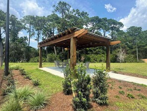 A wooden pergola with a table and chairs underneath, surrounded by a landscaped garden with various shrubs and a clear sky. - Home with Pickleball Courts, Wifi & Beach Essentials (Navarre)