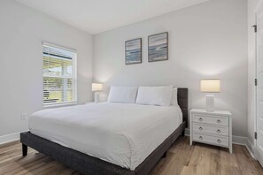 Ein Schlafzimmer mit einem Bett, einem Nachttisch, einer Lampe, einem Fenster mit Jalousien und zwei an der Wand hängenden Bildern.