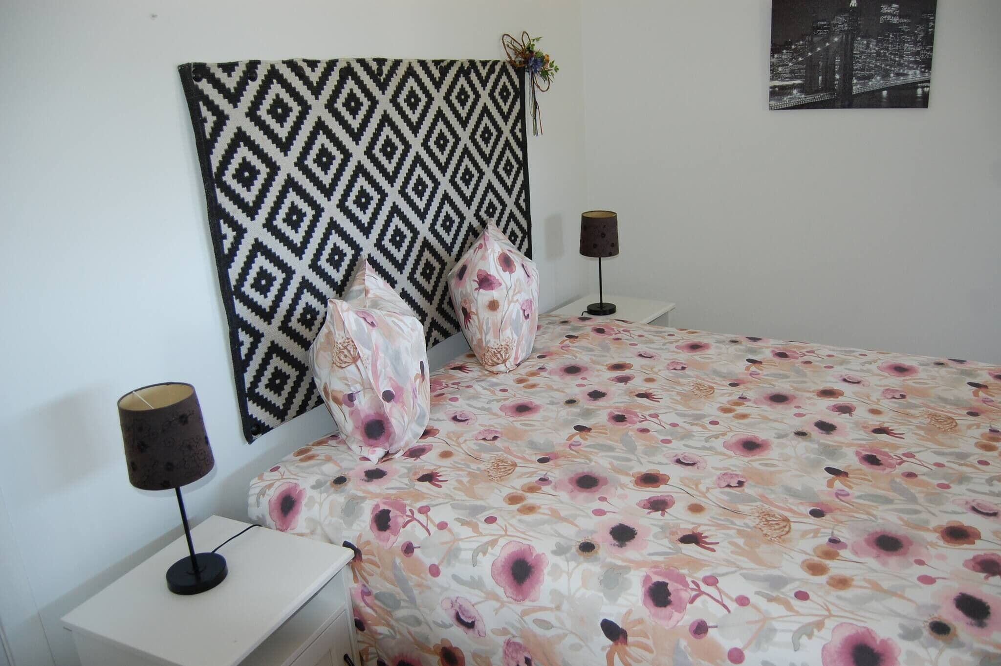 A bedroom with a patterned headboard, floral bedspread, two bedside lamps, and a cityscape view.