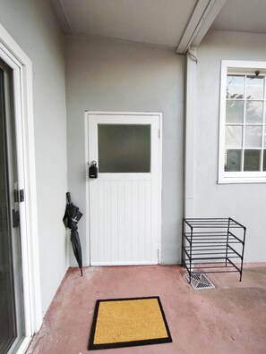 Una puerta blanca con un panel de vidrio esmerilado, un paraguas negro apoyado en la pared, un perchero metálico y un felpudo amarillo sobre un suelo de concreto.