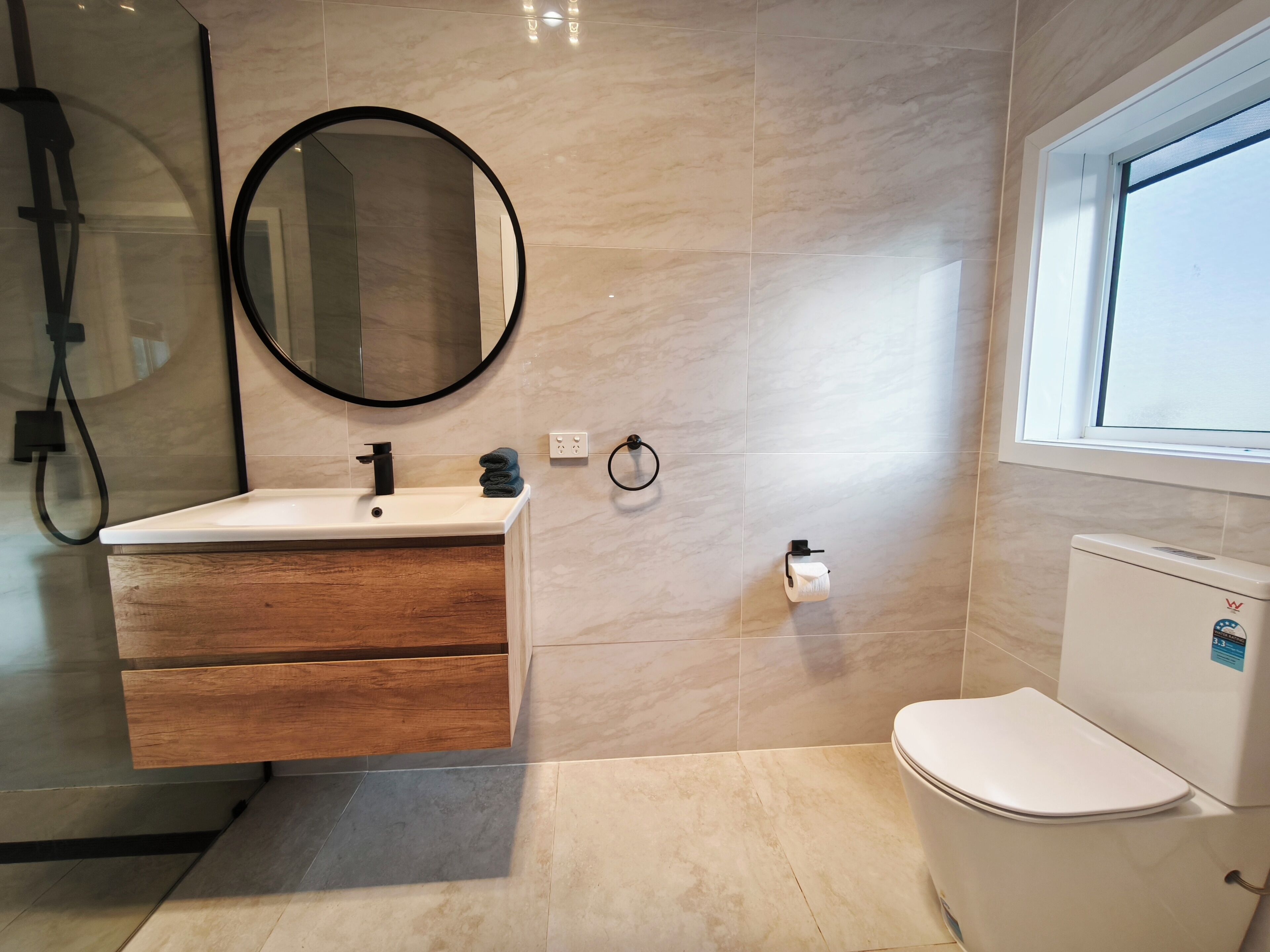 Une salle de bain avec un lavabo blanc, un meuble-lavabo en bois, un miroir rond, des toilettes et une fenêtre.