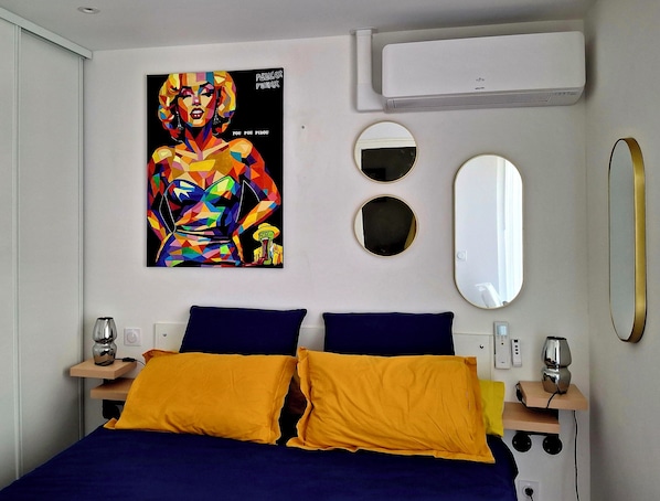 Une chambre à coucher avec un lit, deux oreillers, un climatiseur fixé au mur, deux miroirs ronds et une affiche accrochée au mur.