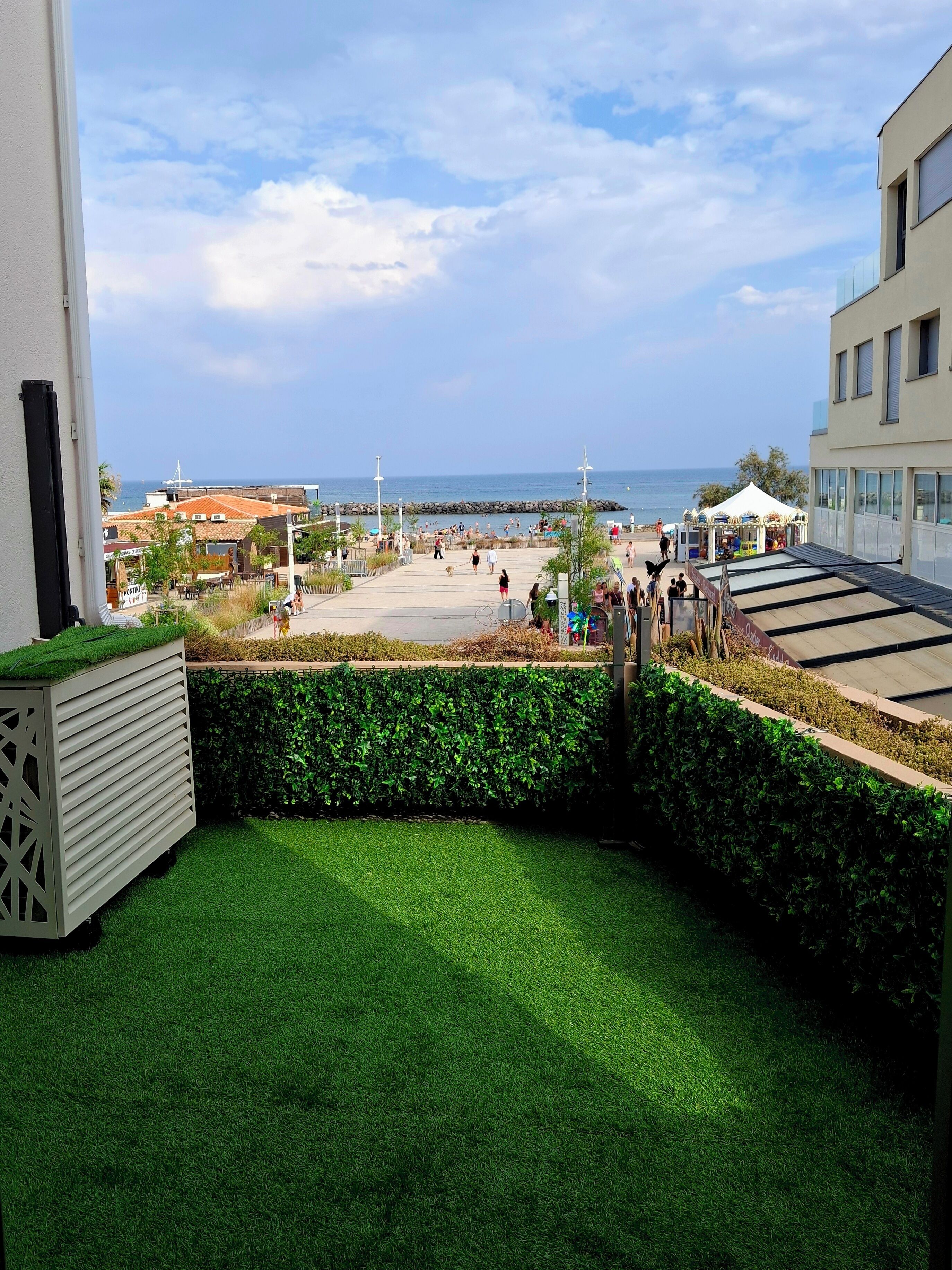 A balcony with artificial grass, a view of the sea, and a promenade with people and shops.