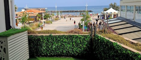 En balkong med konstgräs, utsikt över havet och en strandpromenad med folk och butiker.