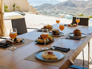 A dining table set with various dishes and drinks, overlooking a mountainous landscape. - YOUTOPIA IOS Luxury Villas & Suite (Ios)