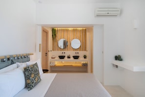A modern bedroom with a bed, a bedside table, a built-in vanity with a mirror, and a wall-mounted air conditioner. - YOUTOPIA IOS Luxury Villas & Suite (Ios)
