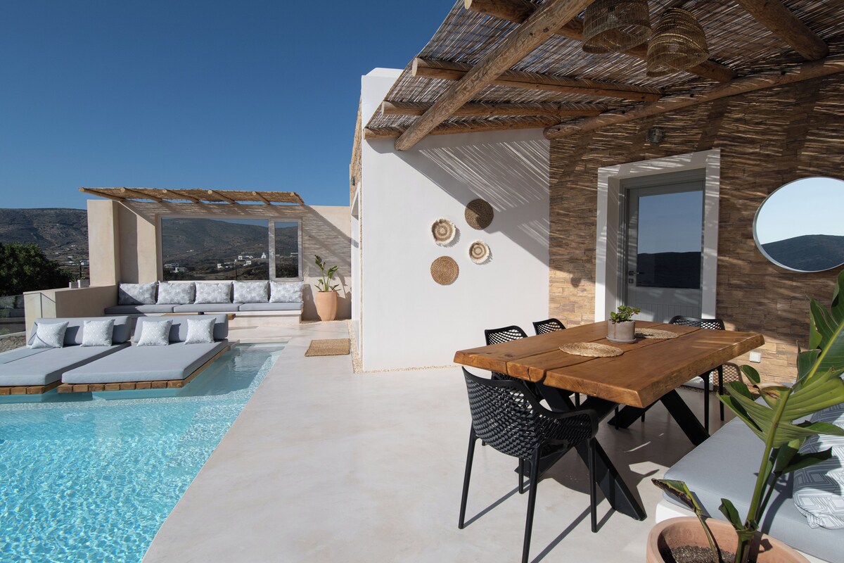 A poolside area with a wooden table and chairs, a lounge with cushions, and a view of the landscape.