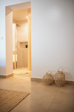 A hallway with a light-colored floor, two wicker baskets, and a door leading to a bathroom. - YOUTOPIA IOS Luxury Villas & Suite (Ios)