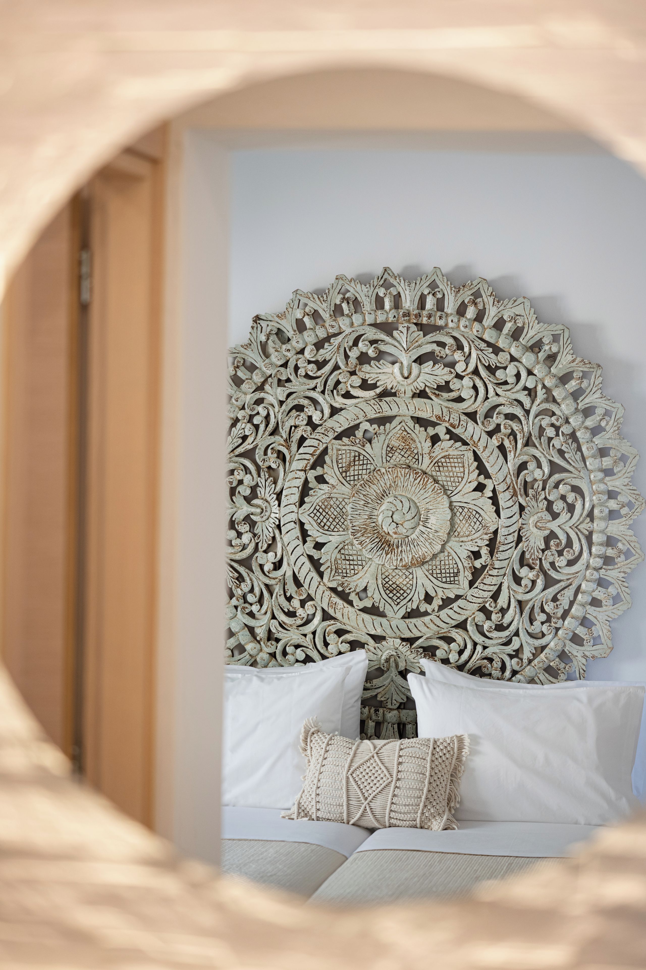 Een slaapkamer met een grote, decoratieve wandversiering, een bed met witte beddengoed en een macramé kussen.