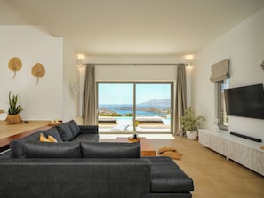 A modern living room with a large sofa, a flat-screen TV, and a view of the sea through large windows. - YOUTOPIA IOS Luxury Villas & Suite (Ios)