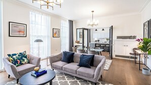 A modern living room with a grey sofa, armchairs, and a coffee table. There is a dining area with a table and chairs, and a kitchen in the background. - Fields in Frame at No. 64 (London)
