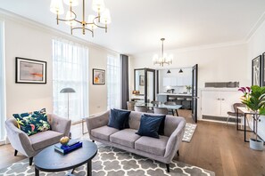 A modern living room with a grey sofa, armchairs, and a coffee table. There is a dining area with a table and chairs, and a kitchen in the background. - Fields in Frame at No. 64 (London)