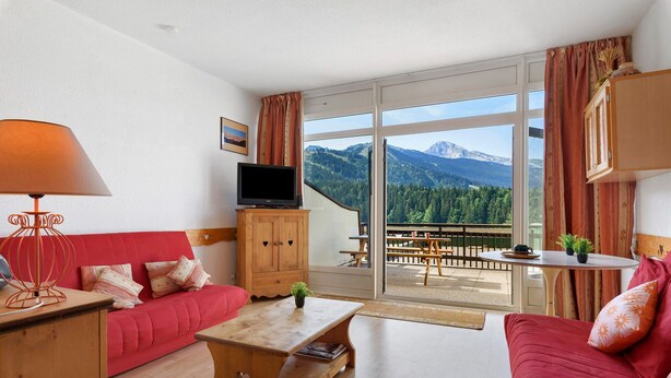 A furnished living room with a red sofa, a wooden coffee table, and a television on a cabinet. Large windows offer a view of mountains and a patio area with outdoor seating. - Superb Mountain View Apartment at the Foot of the Slopes with Large Private Terrace (Villard-de-Lans)