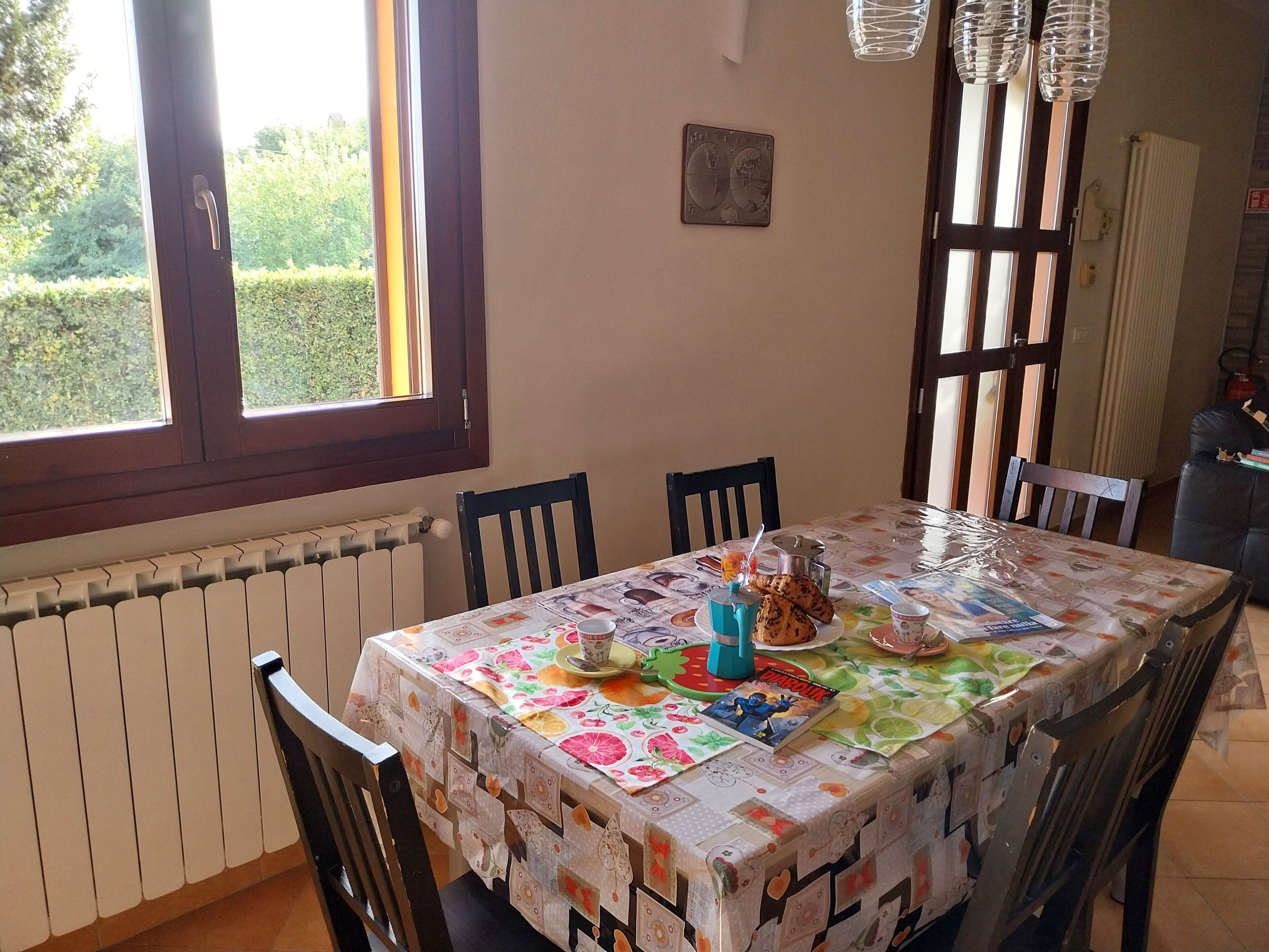Un coin repas avec une table recouverte d’une nappe à motifs, des chaises disposées autour, une fenêtre donnant sur un paysage verdoyant et un radiateur placé sous la fenêtre.