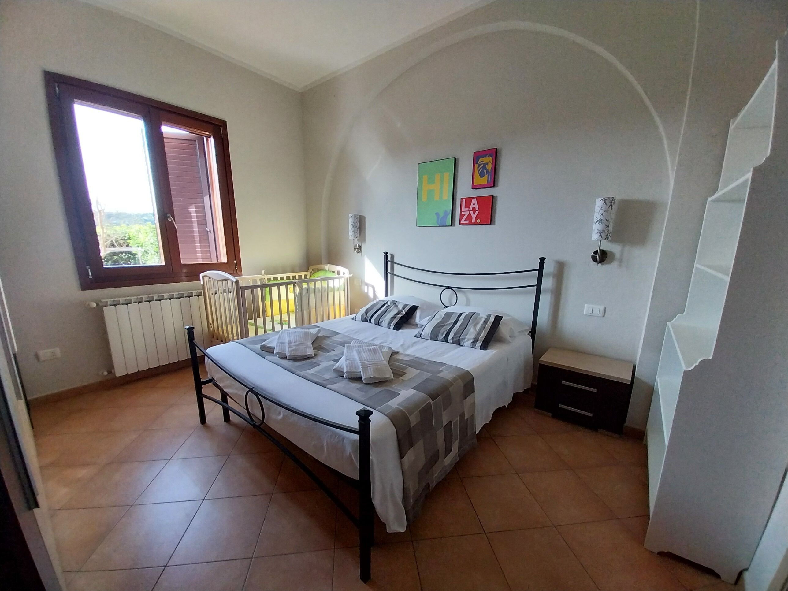 Een slaapkamer met een bed, een raam, een radiator, een ledikant en wanddecoraties.