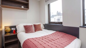 A bedroom with a bed, pillows, a bedside table with a lamp, a window, and a shelf above the bed. - Student Only Crown House (Brighton and Hove)