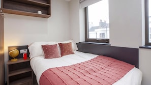 A bedroom with a bed, pillows, a bedside table with a lamp, a window, and a shelf above the bed. - Student Only Crown House (Brighton and Hove)