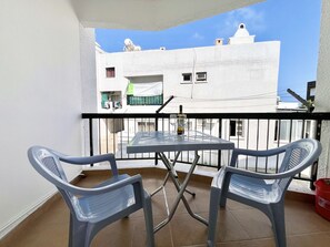 Ein Balkon mit Tisch und zwei Stühlen, einem roten Eimer, und einem weißen Gebäude im Hintergrund.