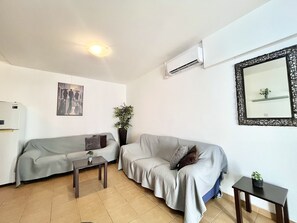 Ein Wohnzimmer mit einer Couch, einem kleinen Tisch, einem Topf mit Pflanze, einer Wandklimaanlage und einem gerahmten Spiegel.