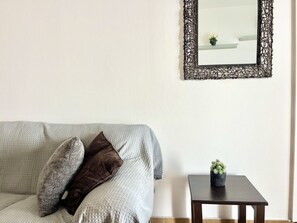 Ein Wohnzimmer mit grauem Sofa, zwei Kissen, einem kleinen Tisch mit Pflanze und einem dekorativen Spiegel an der Wand.