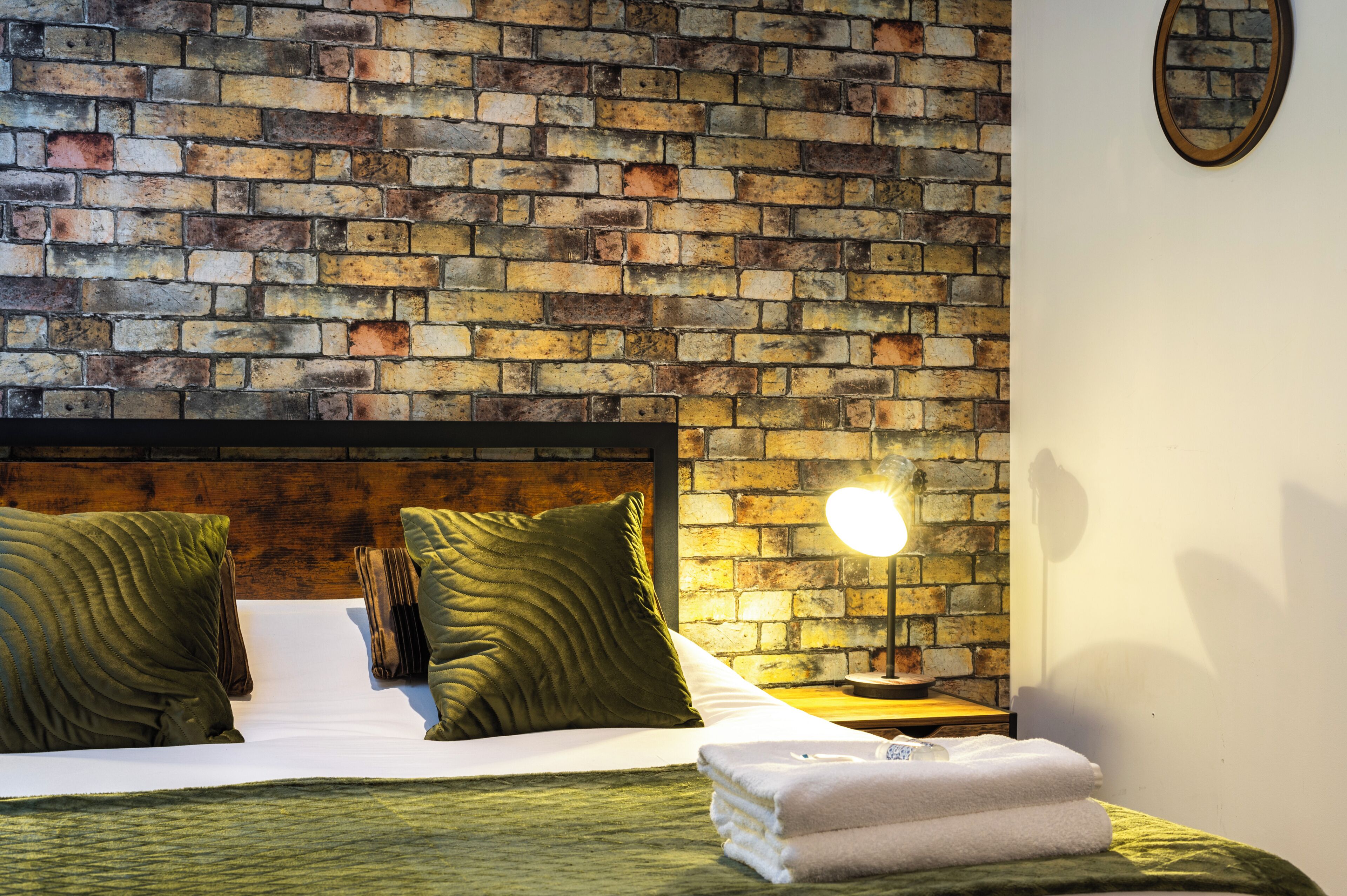 A hotel room with a brick-patterned headboard, a bed with green and white bedding, a bedside lamp, and a stack of white towels.