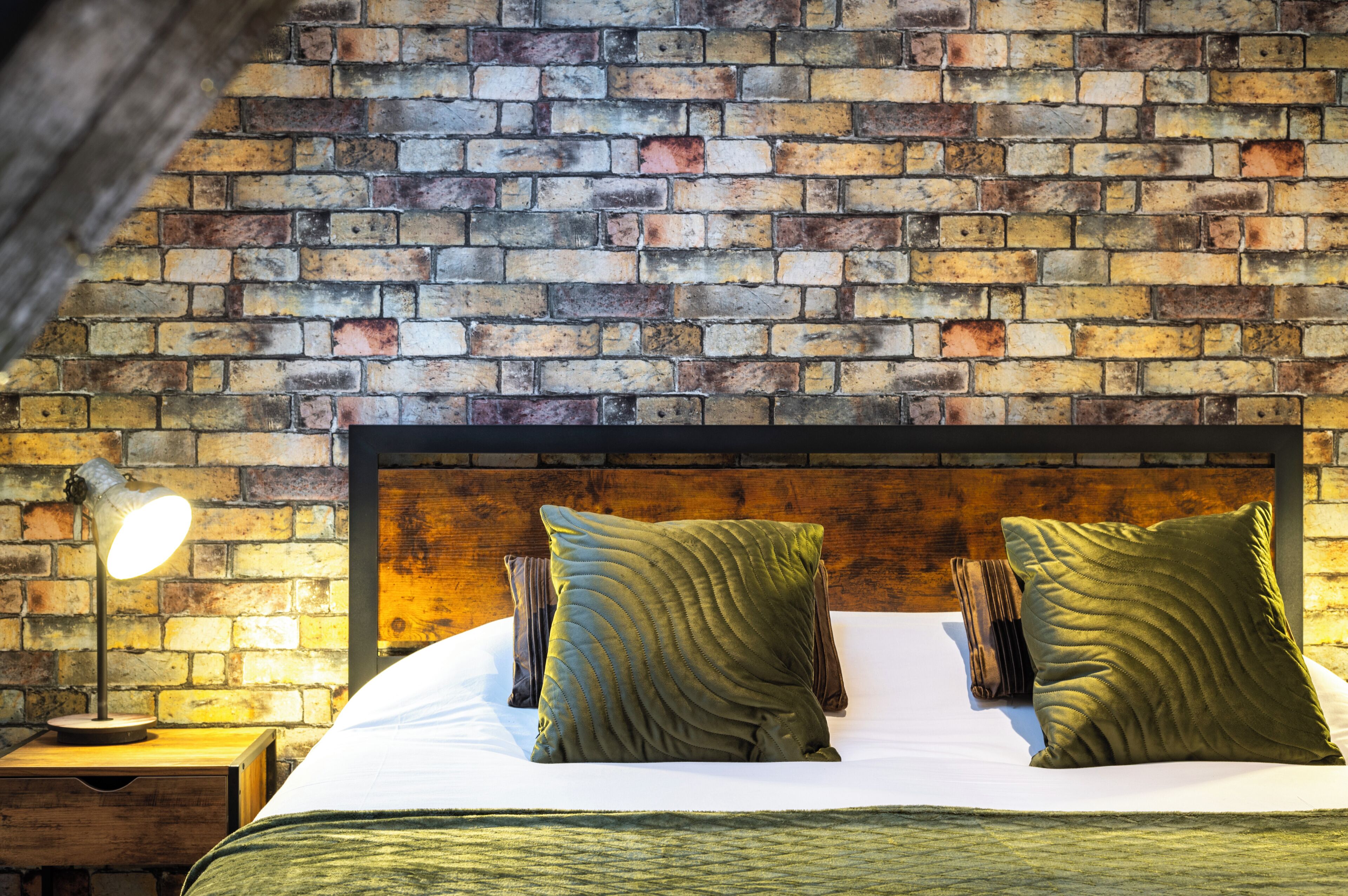 Una habitación con pared de ladrillo, cabecera de madera, cama con almohadas verdes y mesita de noche con lámpara.