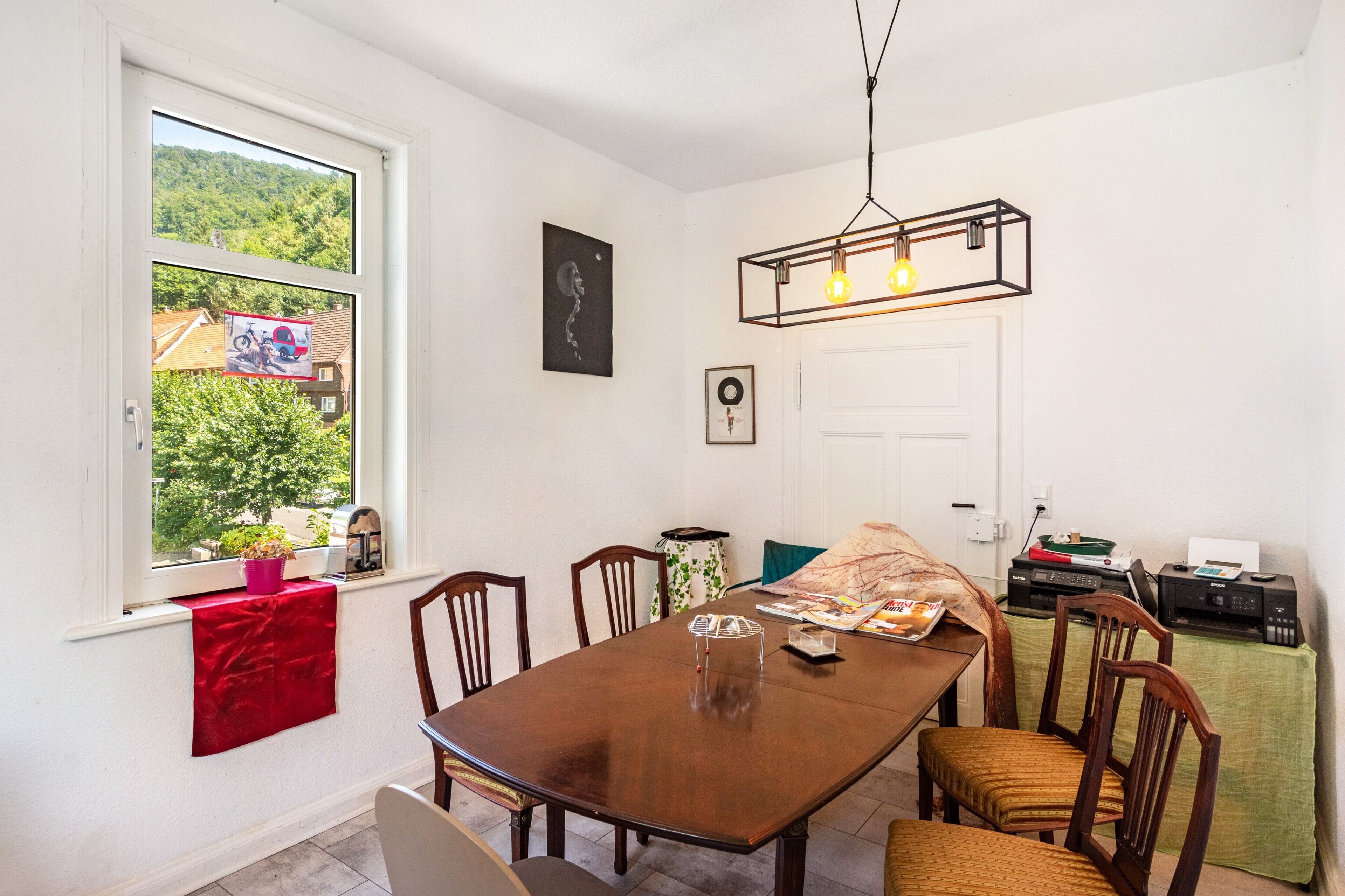 A dining area with a wooden table and chairs, a window with a view of greenery, and a wall-mounted light fixture.