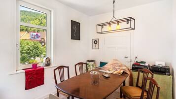 A dining area with a wooden table and chairs, a window with a view of greenery, and a wall-mounted light fixture.