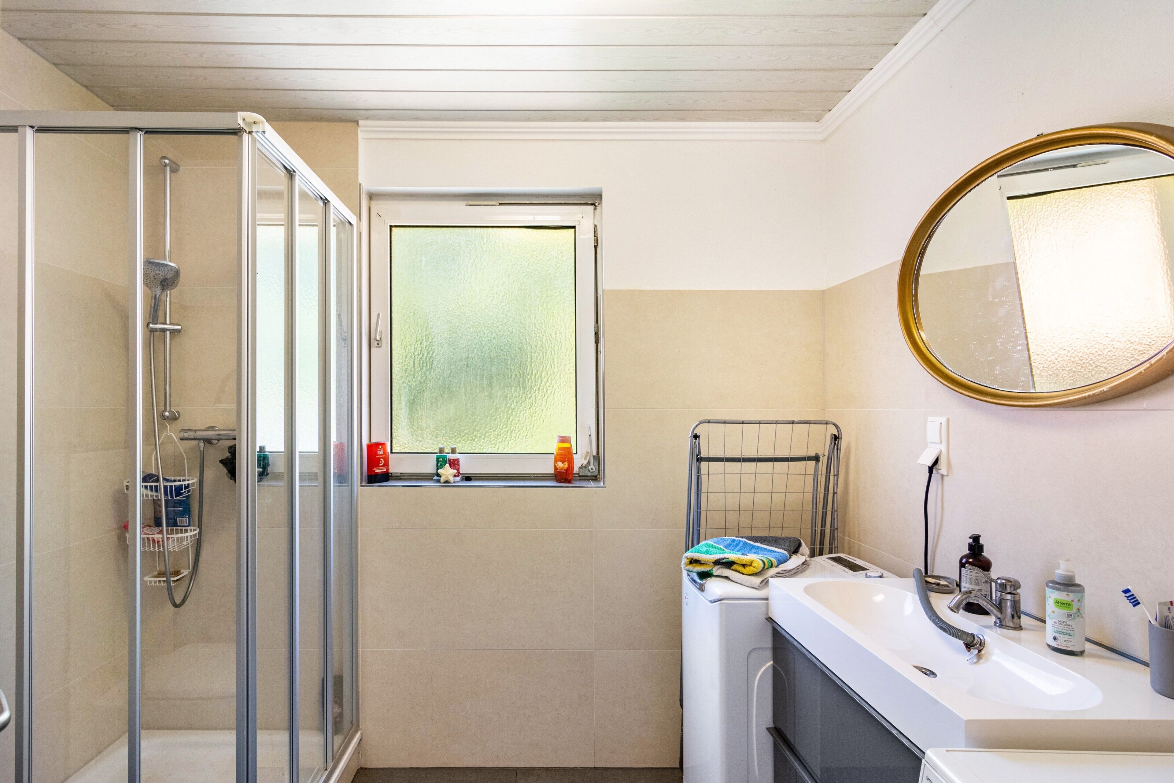 Ein Badezimmer mit Duschkabine aus Glas, einem Fenster mit Blick, einem Waschbecken mit Spiegel und Pflegeprodukten.